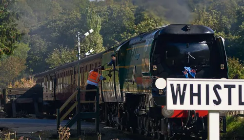 This landscape photograph, taken in the morning during the autumn season of 2018, captures a steam train at the moment of its key exchange on the North Yorkshire Moors Railway in the rural United Kingdom. The scene shows a member of railway staff reaching up from a platform to a train crew member as they exchange the token required for single-track working, which is a classic example of transport operations on heritage railways. The steam train, pulling a row of maroon carriages, is set against a backdrop of green trees, typical of the North Yorkshire Moors area, with signals and a prominent "WHISTLE" sign in the foreground enhancing the authentic rural railway setting.
